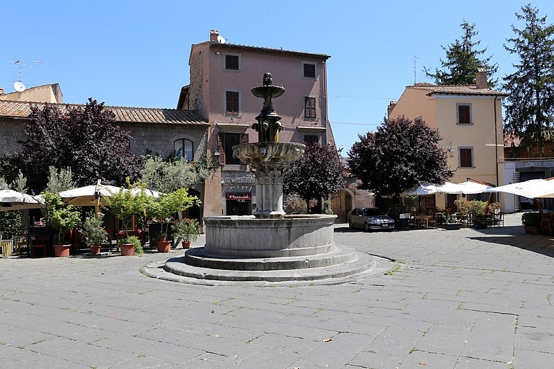 Piazza con al centro una fontana e sullo sfondo edifici e ombrelloni con sedie e tavolini da bar