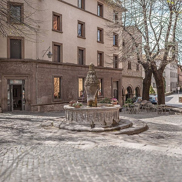 Piazza con al centro un fontana, sullo sfondo edifici e un paio di alberi sulla sinistra preceduti da tavolini e sedie e bar